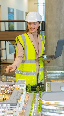 Confident architect in hardhat using laptop while analyzing office complex model at workplace Confident young female architect in hardhat and reflective jacket using laptop and analyzing business office complex architectural model. Woman girl smart designer of buildings working in office.
